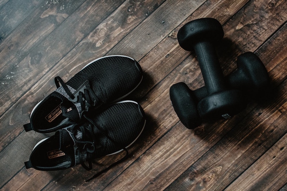 A pair of dumbbells next to a pair of black sneakers on a hard wood floor