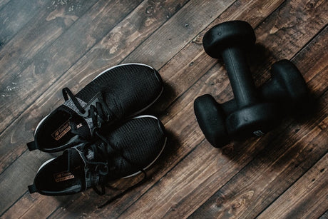 A pair of dumbbells next to a pair of black sneakers on a hard wood floor