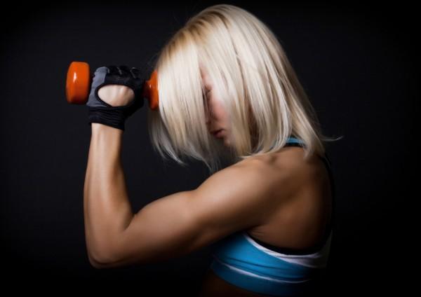 Side view from a blond woman holding up an orange dumbbell